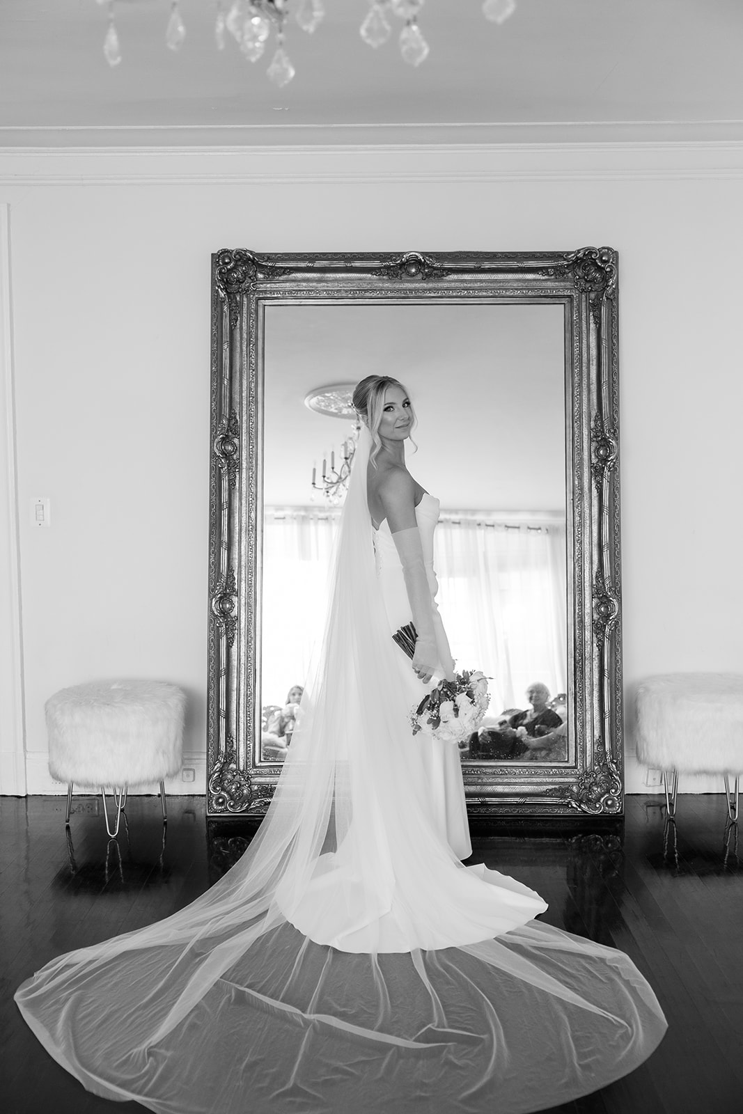 Black and white portrait of the bride with a long flowing veil spread on the floor, posing in front of a mirror.